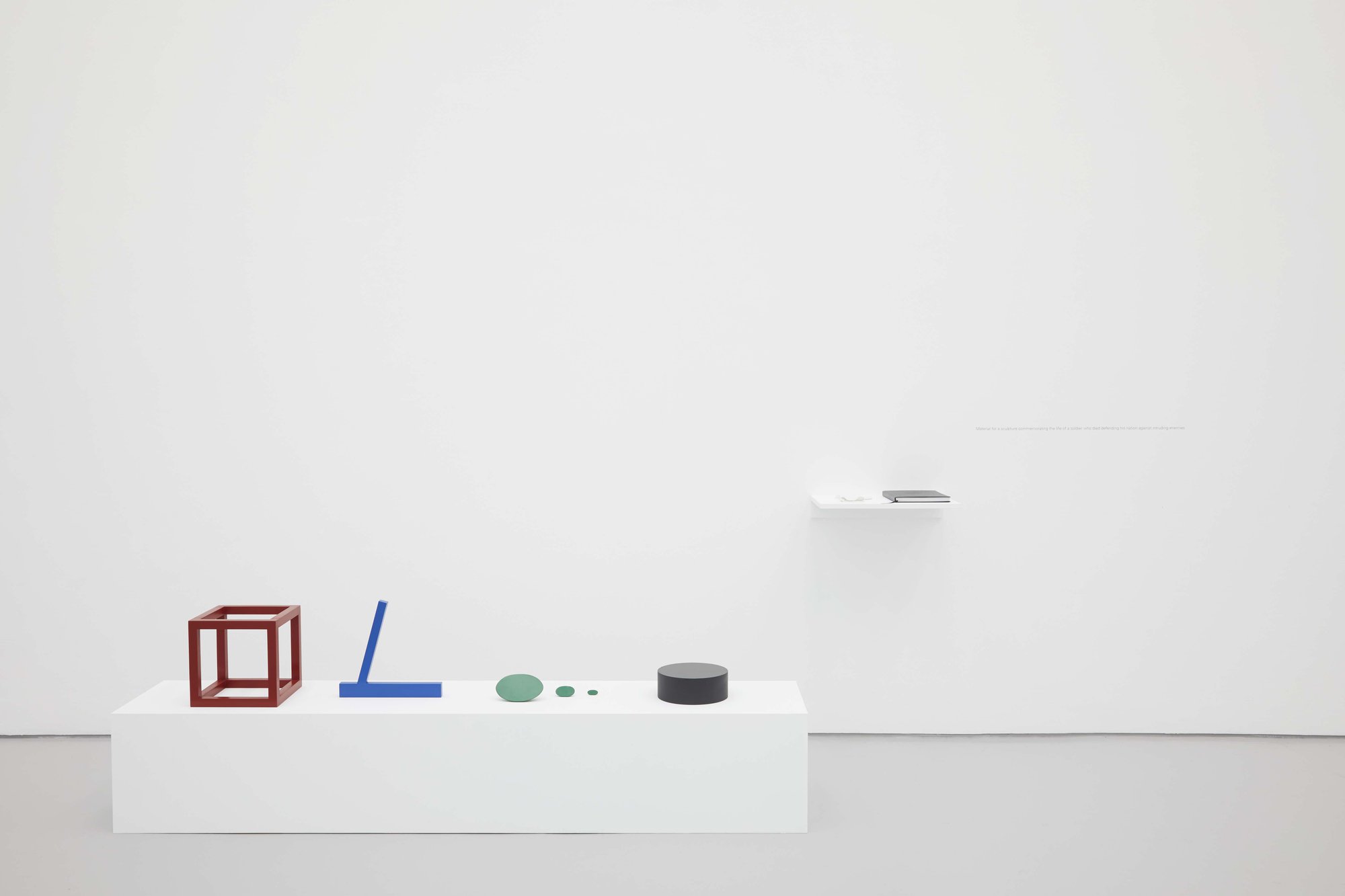 Iman Issa, Material for a sculpture commemorating the life of a soldier who died defending his nation against intruding enemies, four painted wooden sculptures, painted wooden plinth, painted wooden shelf, blank book with four inserts, vinyl text on wall, dimensions variable, 2012